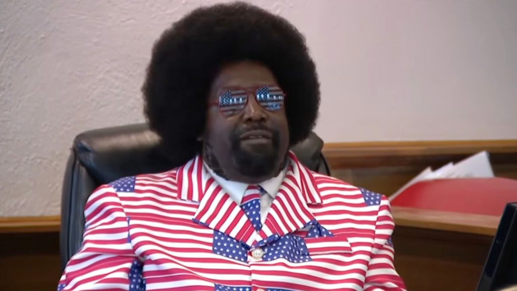 Afroman wearing a red, white, and blue American flag–patterned suit and matching star-spangled tie and sunglasses, seated indoors.