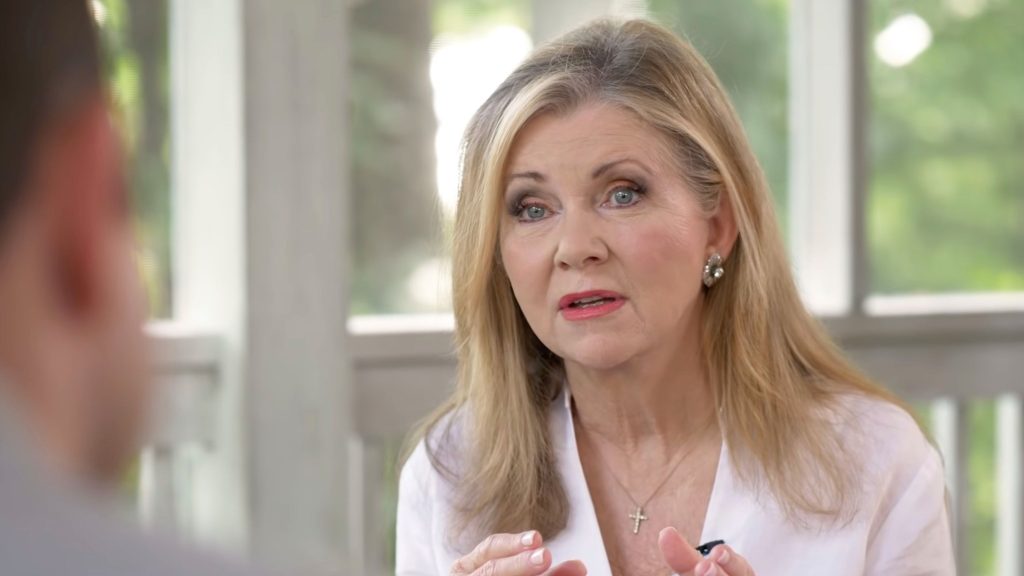 Blackburn with long blonde hair and pearl earrings speaking expressively, wearing a white blouse and cross necklace in a bright room.