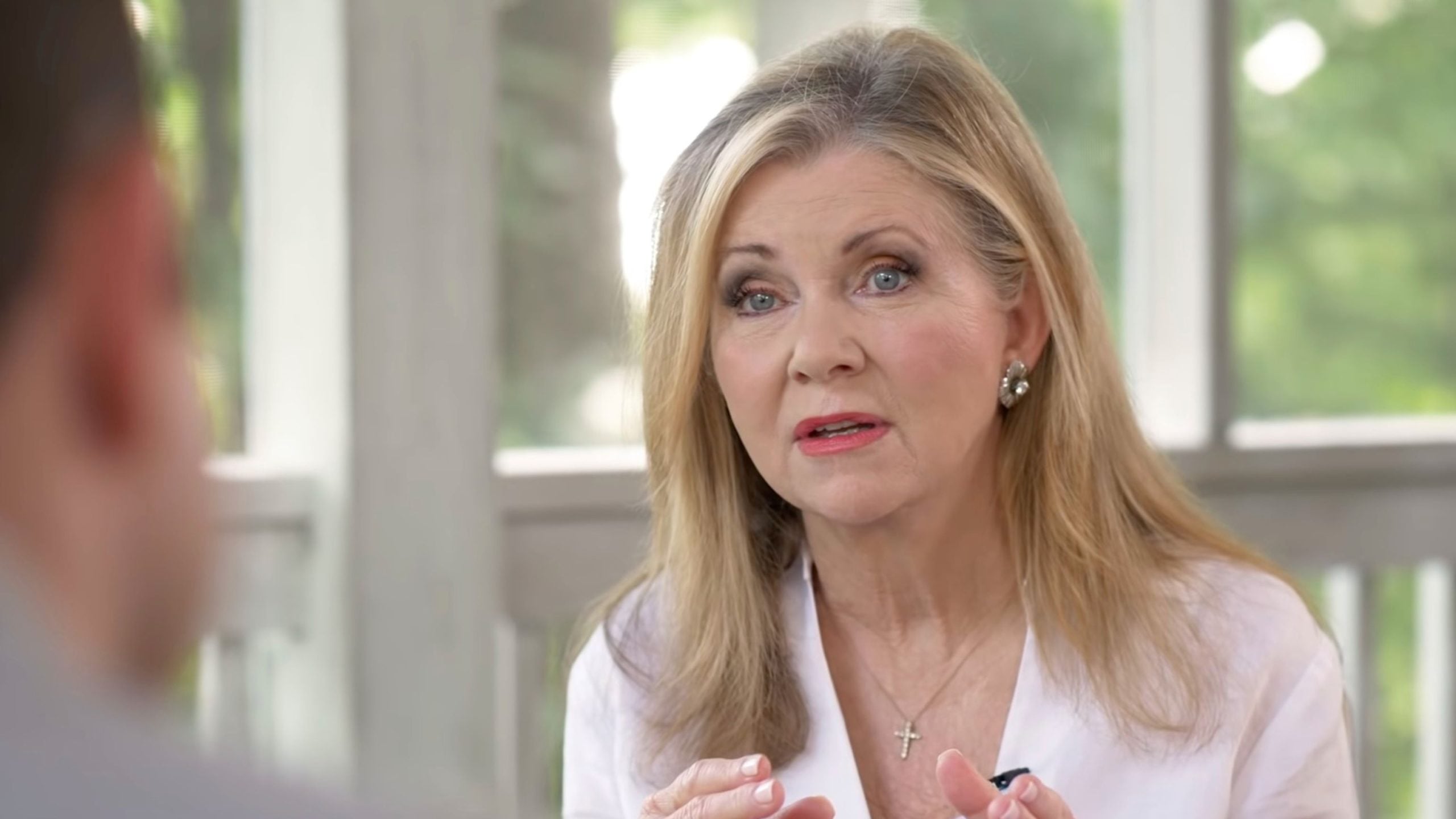 Blackburn with long blonde hair and pearl earrings speaking expressively, wearing a white blouse and cross necklace in a bright room.
