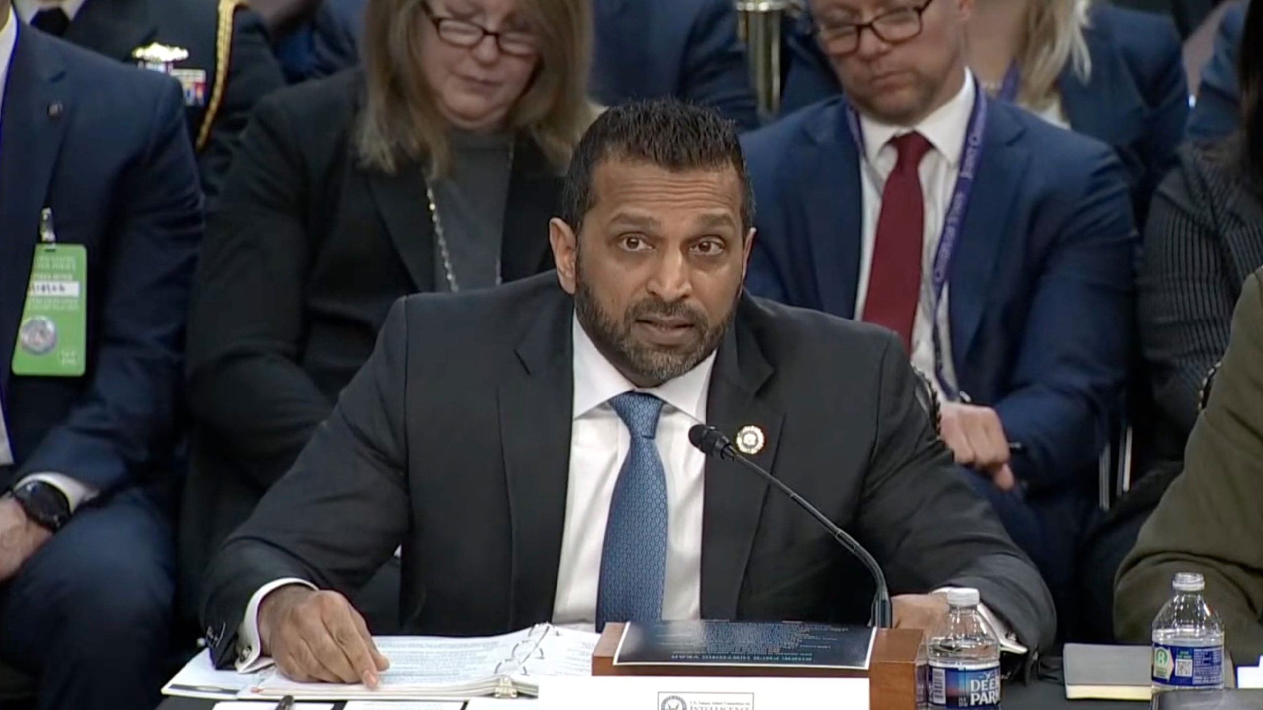 Kash Patel in a dark suit and blue tie speaking into a microphone at a hearing table, papers and water bottles in front of him.