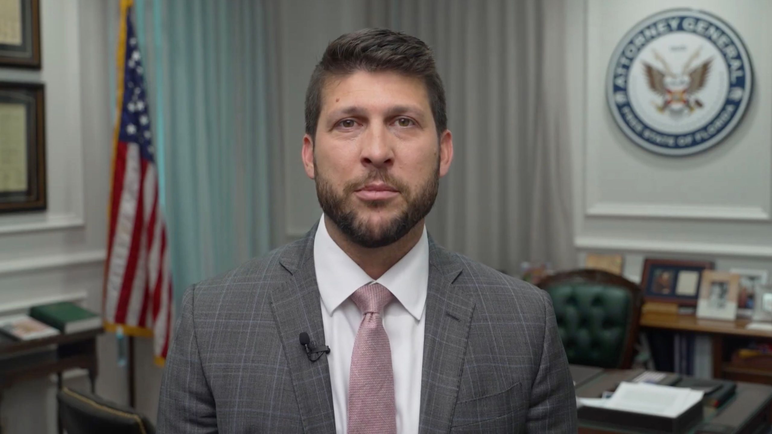 James Uthmeier in a gray plaid suit and pink tie standing in an office with US flag and government seal visible behind him.