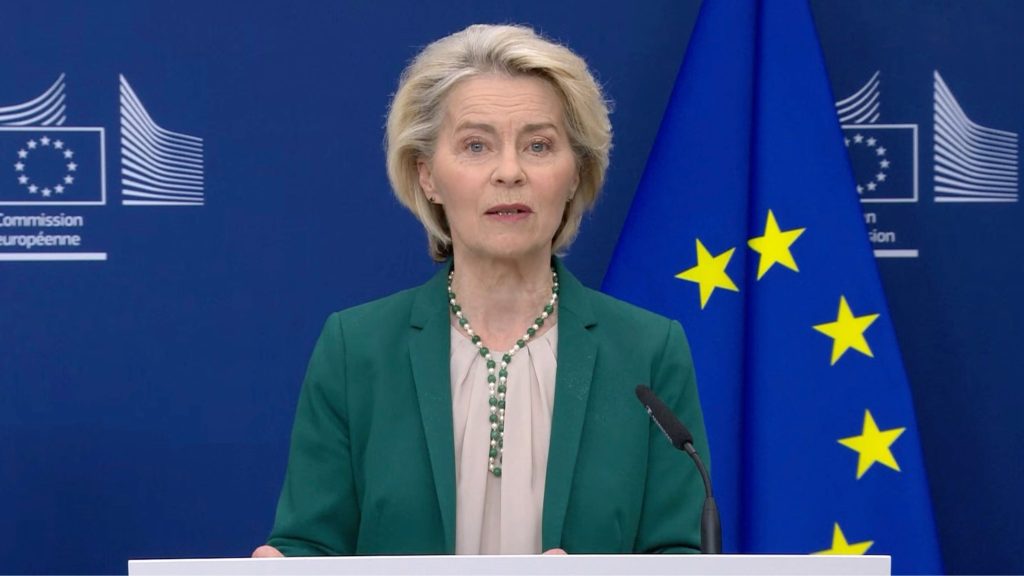 Ursula von der Leyen in a green blazer speaking at a podium with an EU flag and blue backdrop behind her.