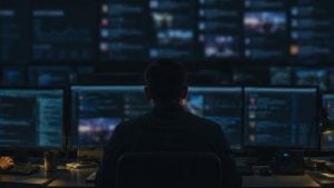 Person seen from behind sitting at a desk with multiple blurred monitors displaying data and feeds in a dark control room.