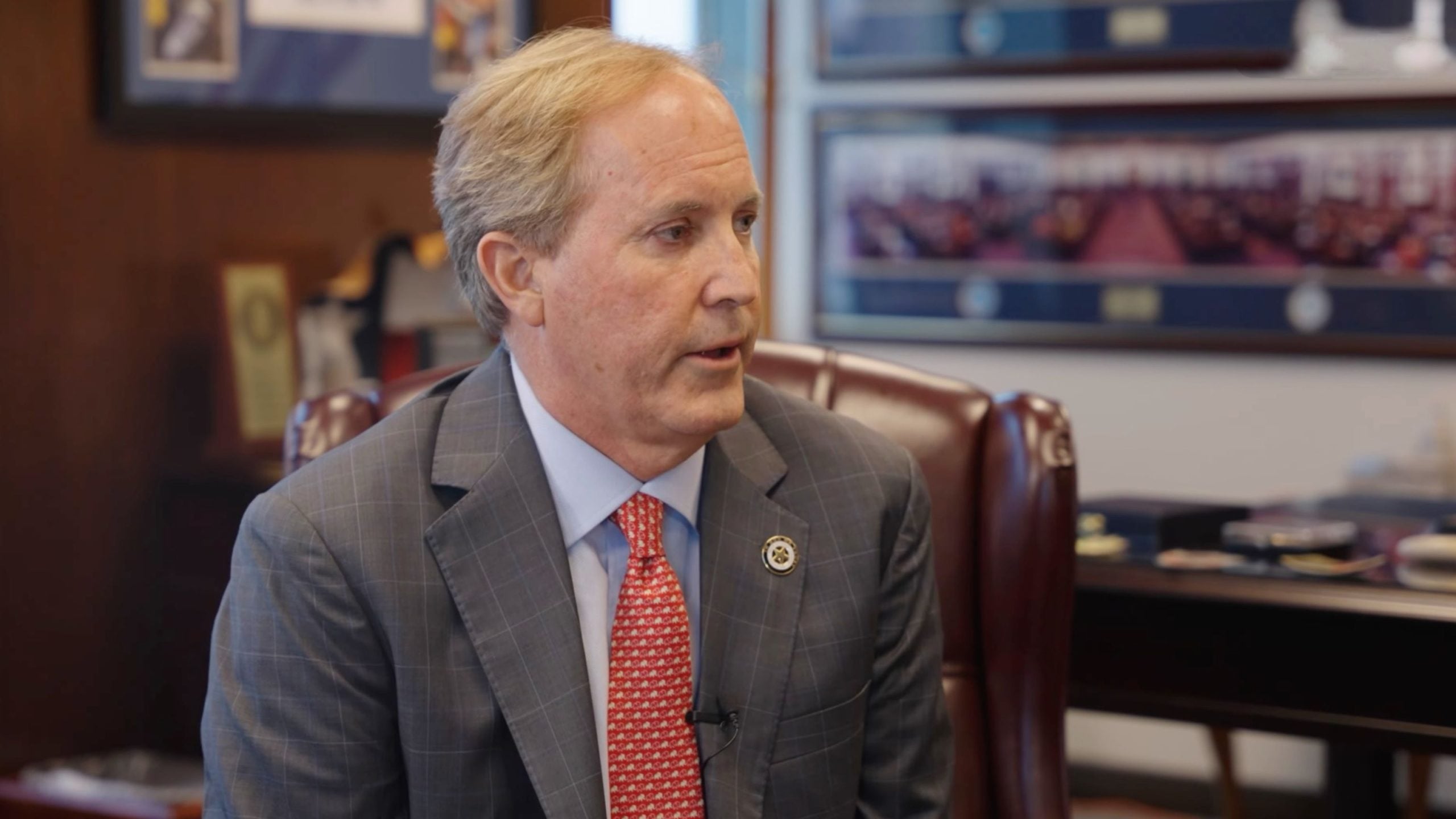 Paxton in a gray checked suit and red patterned tie sitting in an office chair, facing right during a conversation.