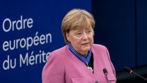 Merkel wearing a pink blazer and a blue-and-gold medal ribbon speaking at a podium with a blue backdrop showing partial French text.