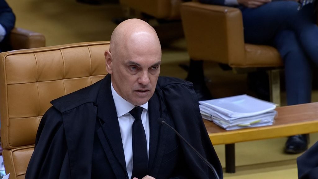 Moraes in a black robe and tie speaking into a microphone while seated at a desk with stacked documents.