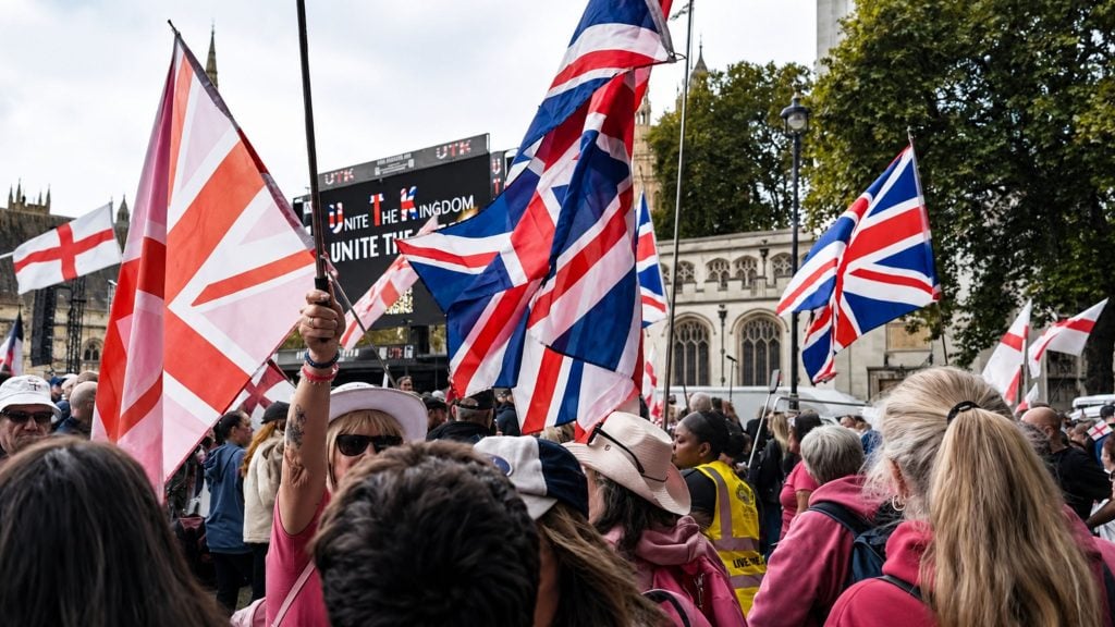 Crowd of people waving multiple Union Jack and St George flags outside a historic building during a rally.