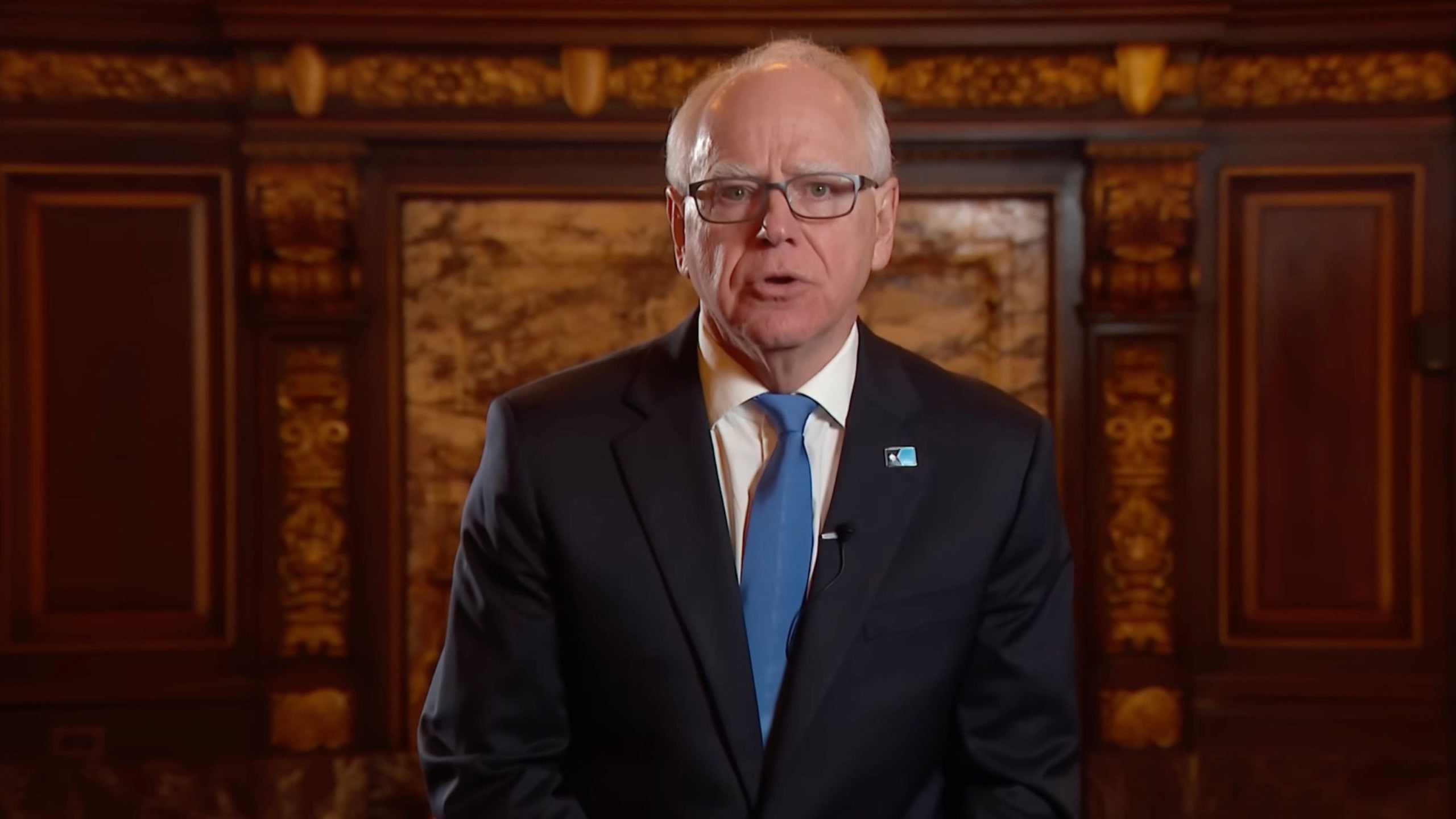 Walz in a dark suit and blue tie speaking to camera against an ornate wood and marble backdrop, wearing glasses and a lapel pin