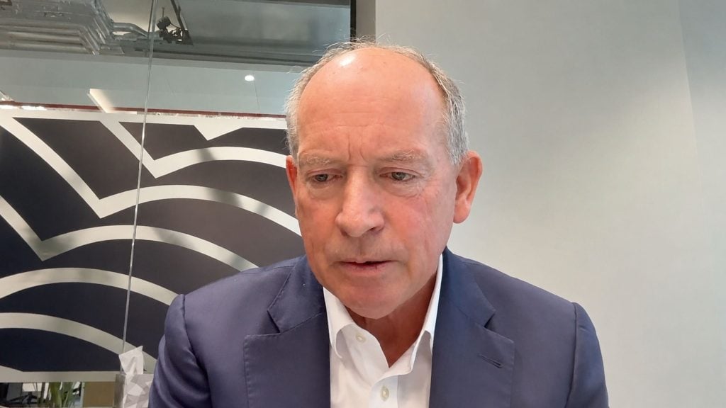 Ian Cheshire wearing a navy blazer and white shirt, looking down in a modern office with patterned glass behind him.