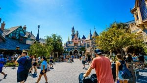 Crowded theme-park street leading to a pastel fairy-tale castle with blue turrets, surrounded by medieval-style buildings and trees.