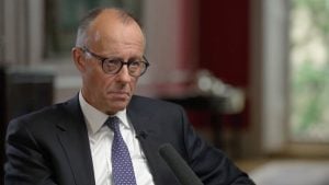 Merz in a dark suit, white shirt and patterned tie wearing round glasses and listening intently during an interview.
