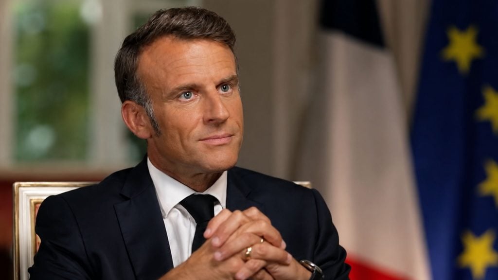 Macron in a dark suit and tie sitting with hands clasped, looking slightly upward against a blurred background with flags.