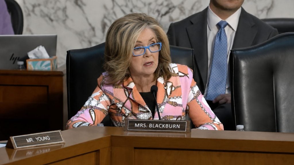 Blackburn with shoulder-length blonde hair and blue glasses speaking into a microphone at a wooden hearing desk labeled "Mrs. Blackburn," wearing a patterned pink and orange jacket.