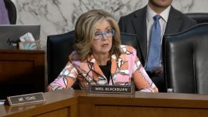 Blackburn with shoulder-length blonde hair and blue glasses speaking into a microphone at a wooden hearing desk labeled "Mrs. Blackburn," wearing a patterned pink and orange jacket.