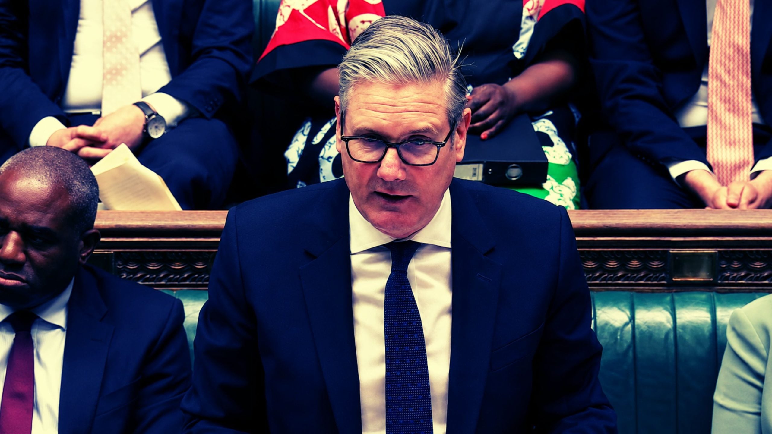 Starmer in glasses and a dark suit speaking in a formal chamber, seated on green leather benches with others behind him.