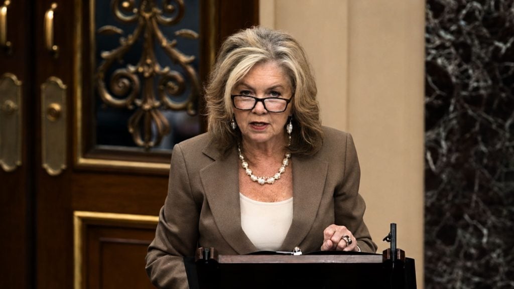 Blackburn with shoulder-length blonde hair and glasses speaking at a podium, wearing a brown blazer and pearl necklace in a formal room