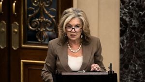 Blackburn with shoulder-length blonde hair and glasses speaking at a podium, wearing a brown blazer and pearl necklace in a formal room