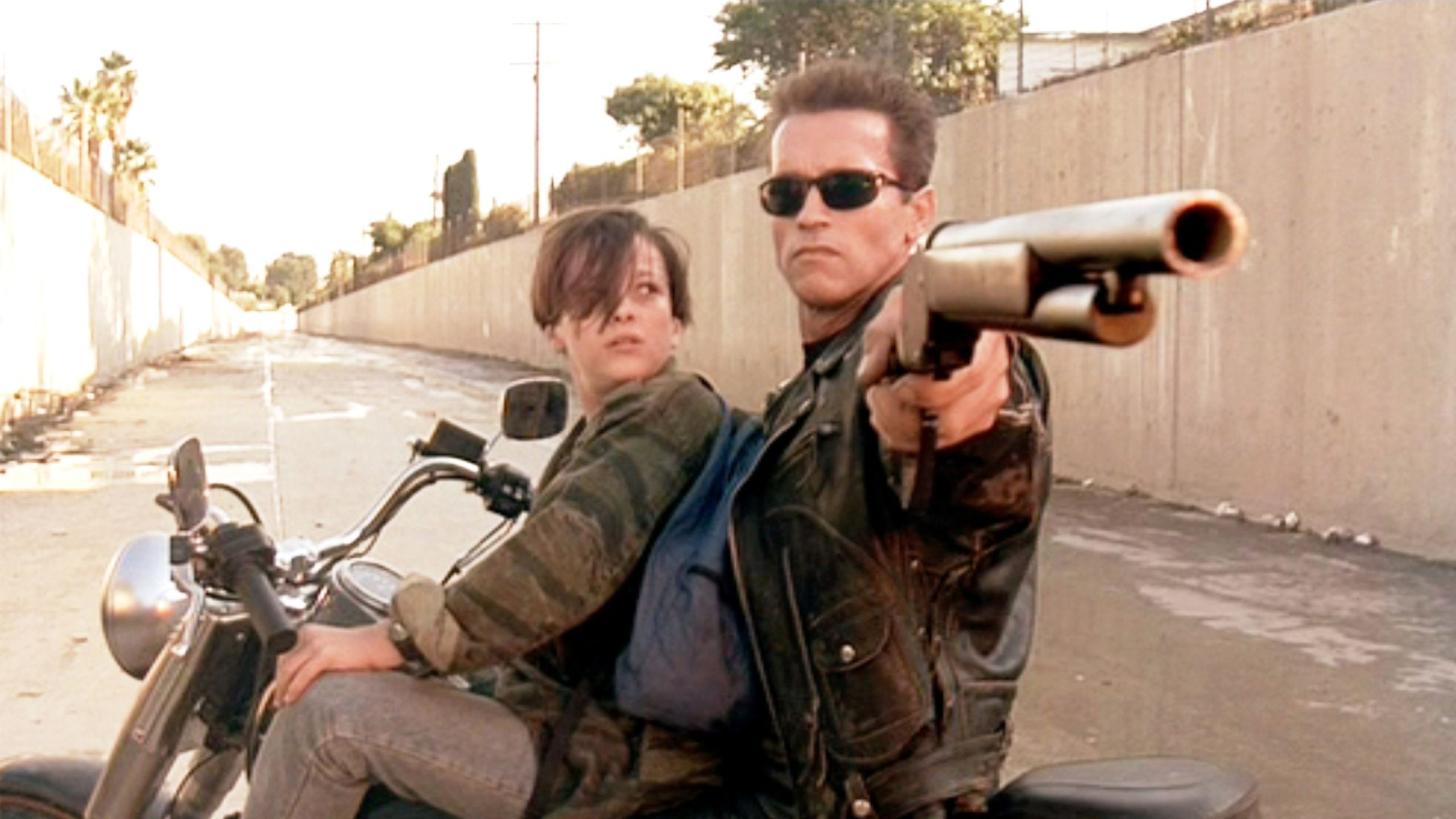 The Terminator on a motorcycle points a shotgun while a woman sits behind him in a concrete channel
