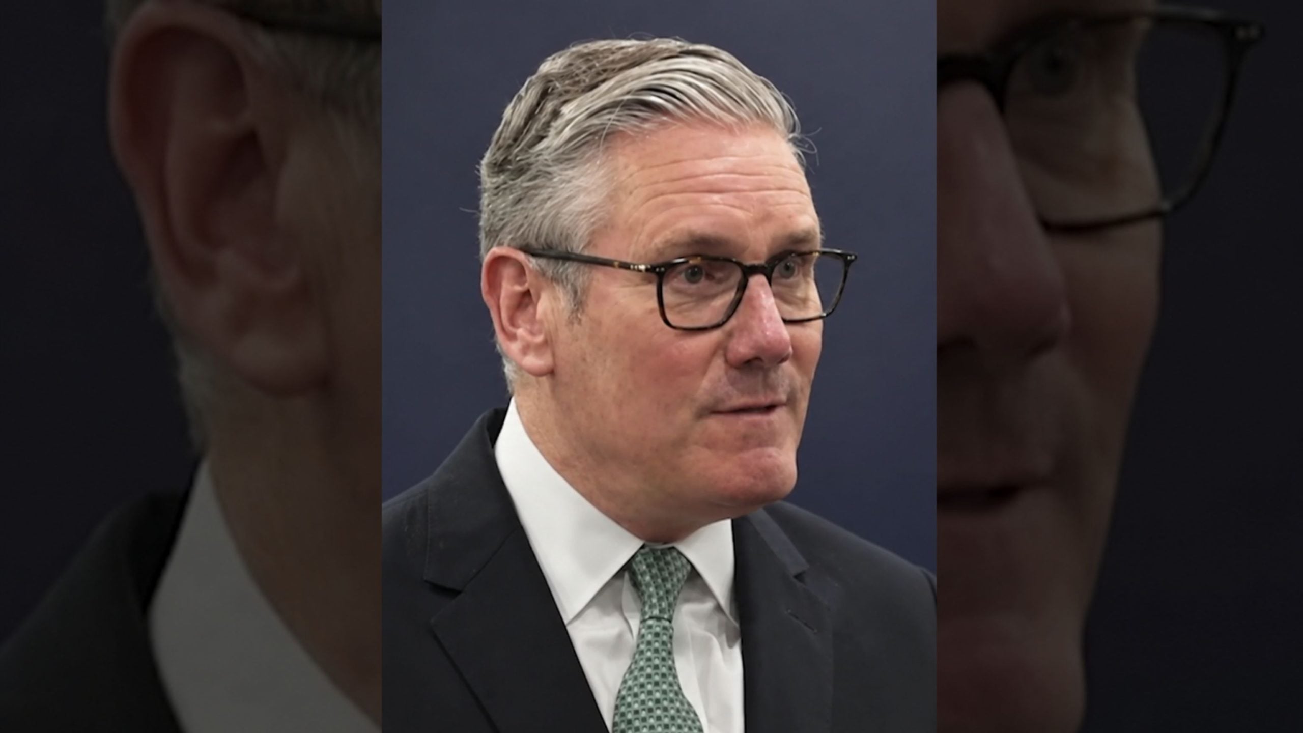 Starmer with gray swept-back hair and glasses wearing a dark suit, white shirt and patterned green tie against a dark background.