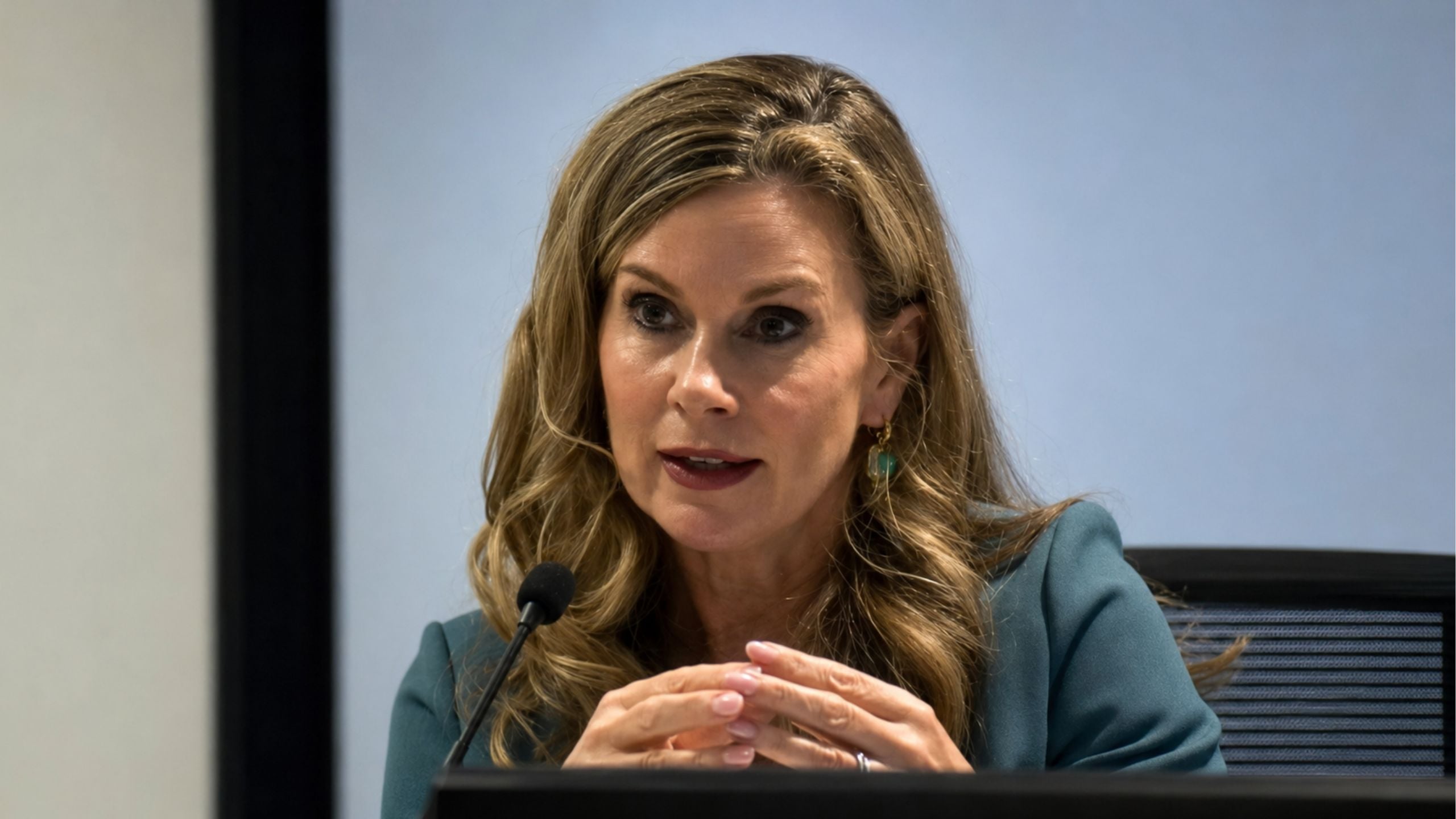 Inman Grant speaking into a microphone at a conference table, wearing a teal blazer and earrings