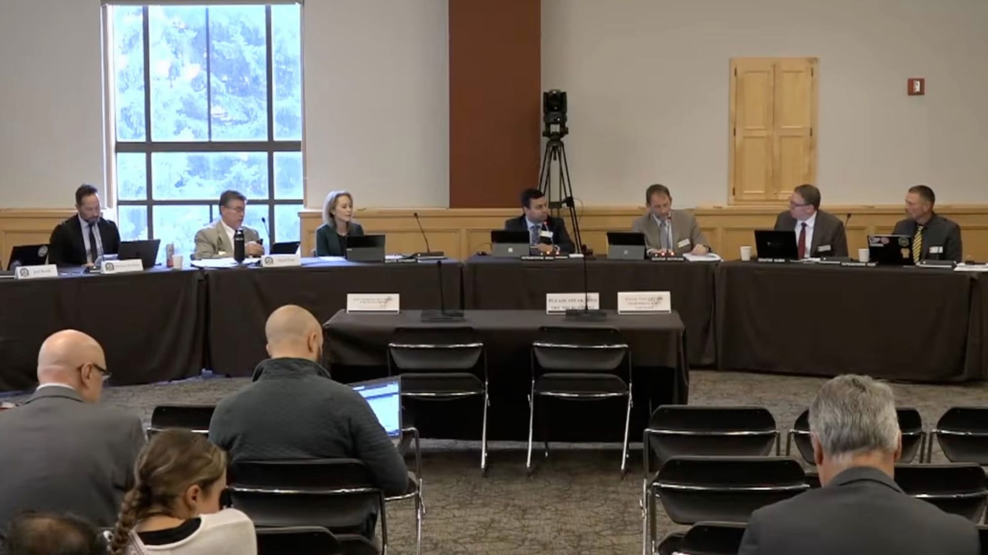 Committee members sit behind a table with laptops during a government hearing in a conference room.