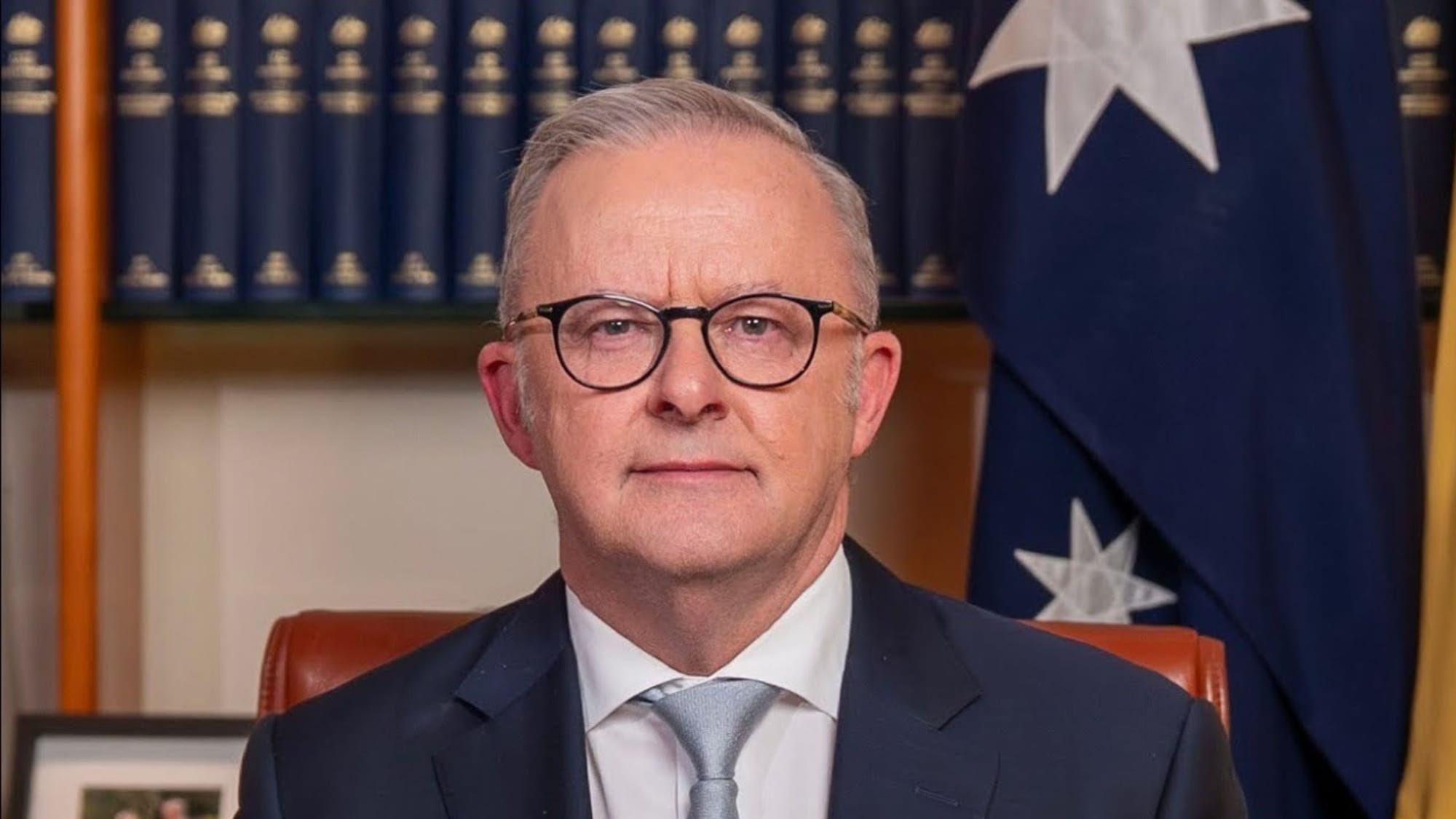 Australian Prime Minister Anthony Albanese wearing glasses and a suit in front of a bookshelf and the Australian flag.