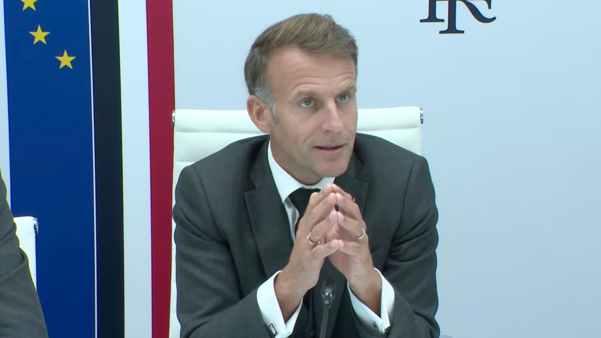 Emmanuel Macron speaks with his hands clasped while seated at a meeting in front of European Union and French flags.