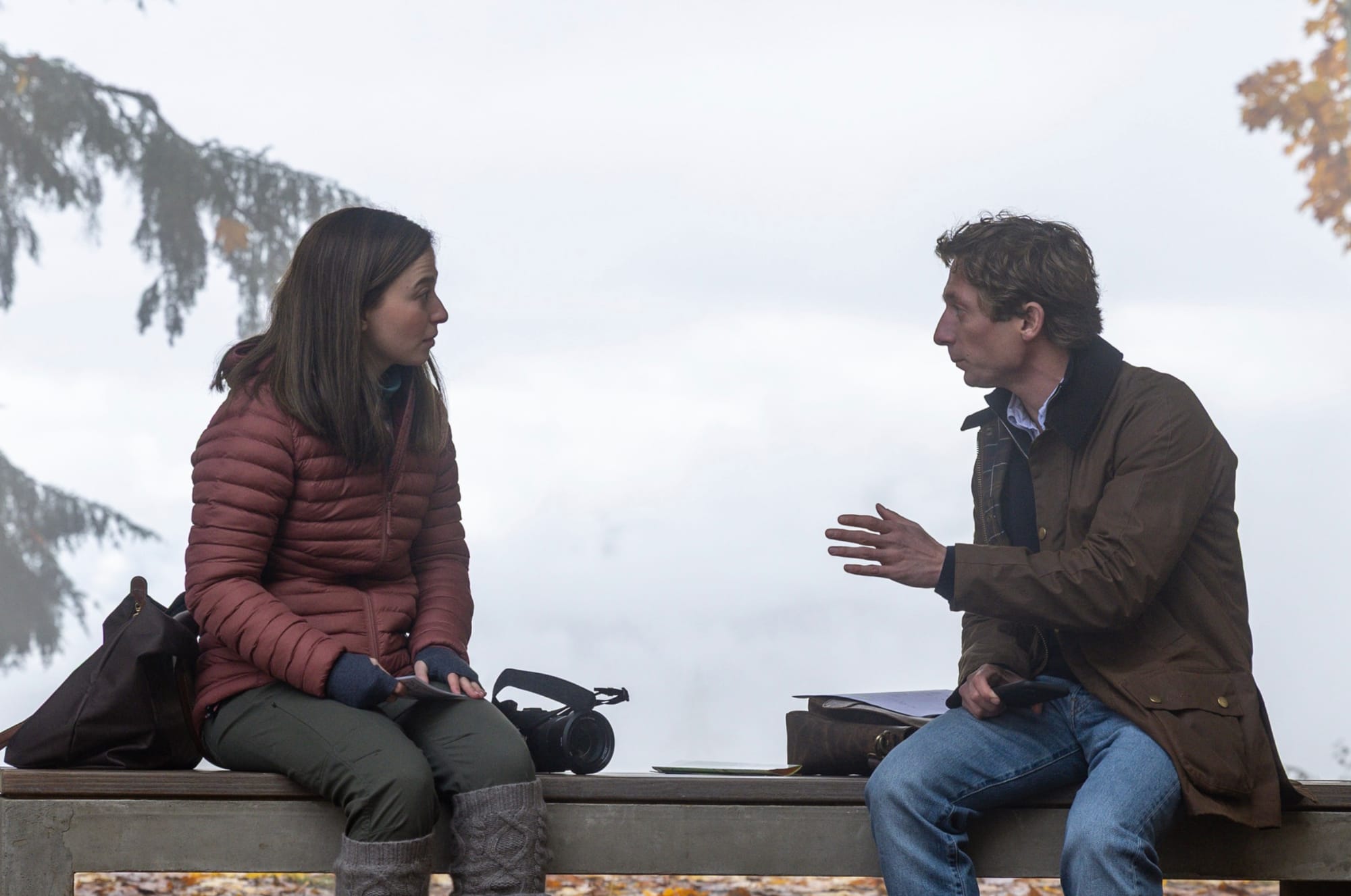 A man and a woman in outdoor jackets sit on a bench outdoors and talk on an overcast day.